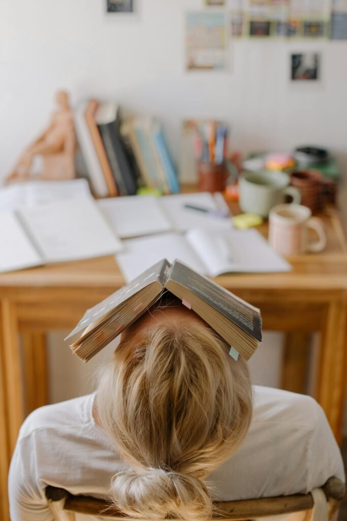 pexels-photo-4769486-4769486 Student feeling stress and exhaustion while studying at a cluttered desk with an open book on their head.